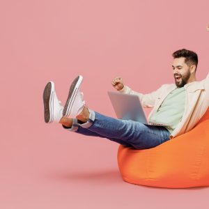 Full body young eacited overjoyed cool happy man in trendy jacket shirt sit in bag chair hold use work on laptop pc computer do winner gesture isolated on plain pastel light pink background studio